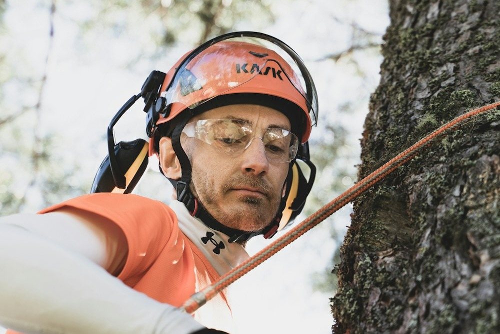 A climber in a helmet and safety glasses roped in against a trunk, reaching for a limb