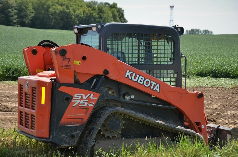 A compact track loader parked at the edge of a field beside a tree line